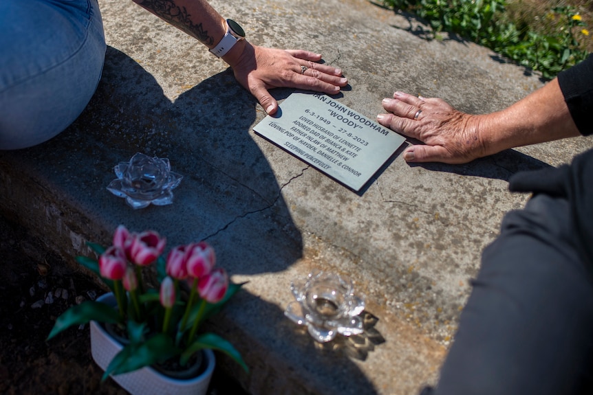 Two hands rest on the silver plaque that reads "Ian John Woodham" alongside pink flowers and crystal ornaments.