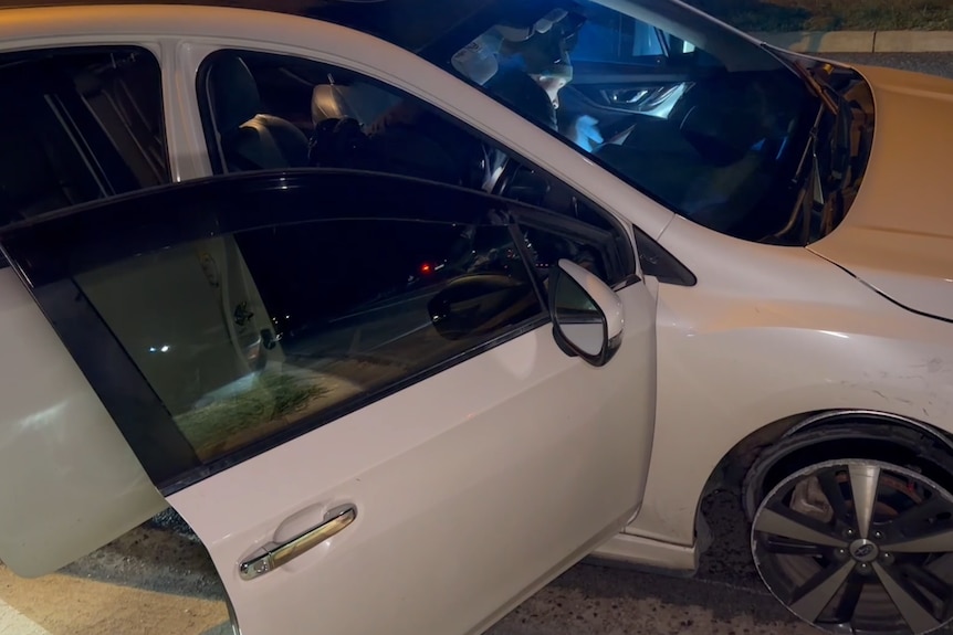 A person leans into the front seats of a white car, shining a torch at something in the interior at night.