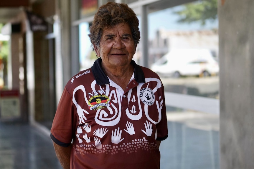 A woman in a patterned red shirt standing on a footpath in front of a window.