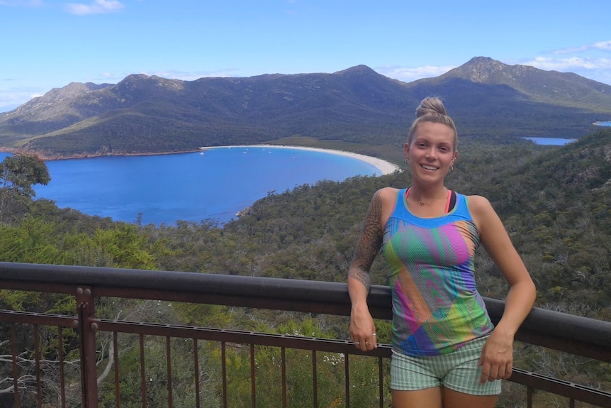 A young woman with blonde hair and a colourful rainbow singlet and shorts stands at a lookout with ocean bay in background