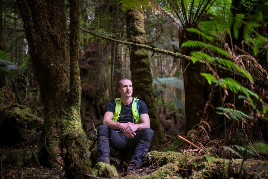 A man wearing long pants and a hi-vis vest with short, shaved hair sits on a log in a moss-covered forest
