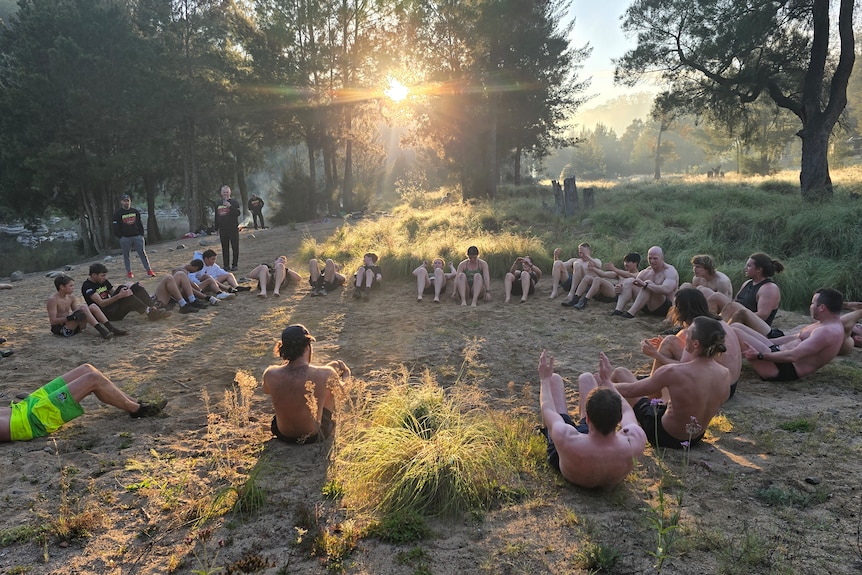 A group of about 30 people sit in a circle on the bank of a river.