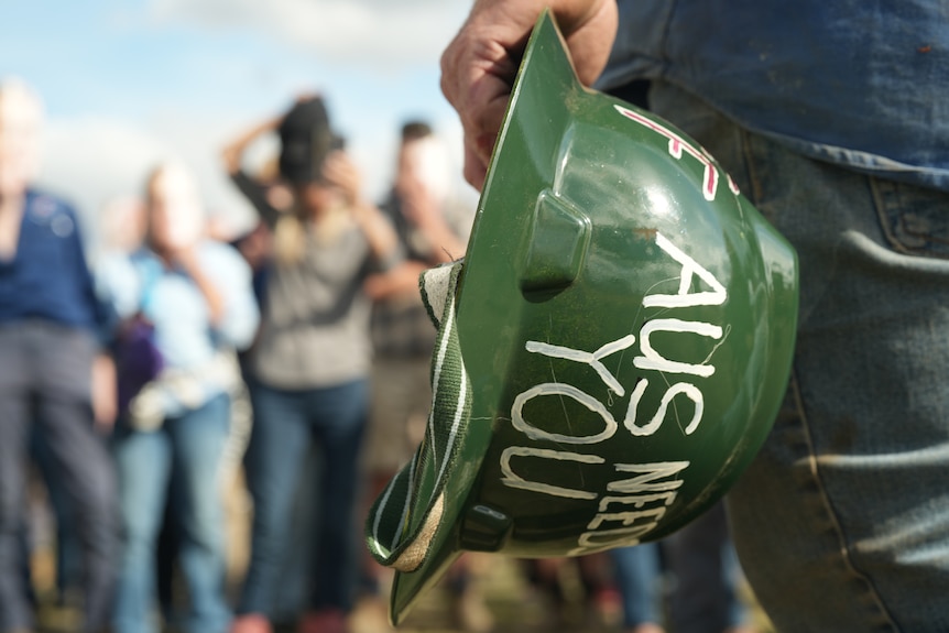 Close up man's hand holding green helmet reading "Aus needs you" in white letters, blurry crowd in background