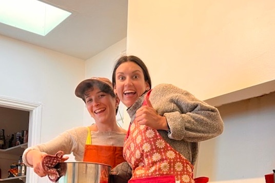 Penny Bradley and another young woman smile while holding a heavy pot and wearing aprons.