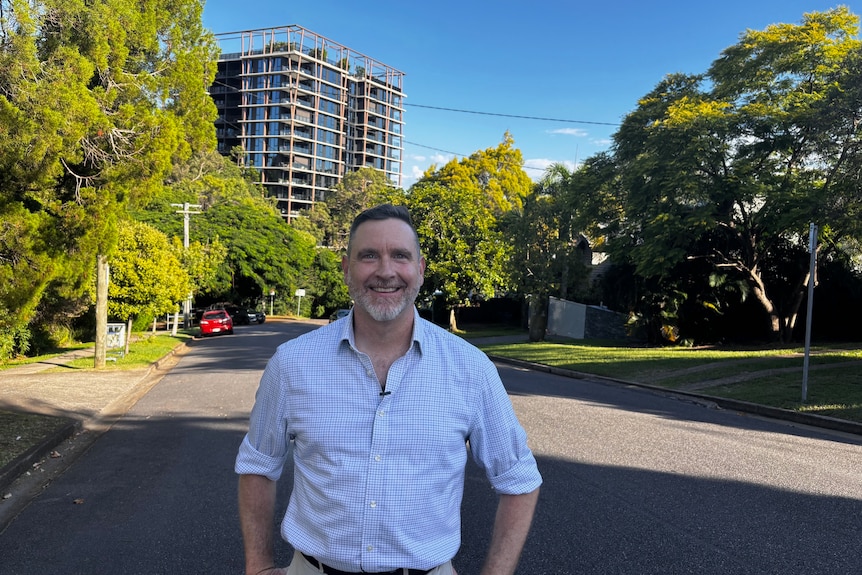 A man in a checked shirt stands on an urban street with a high-rise apartment in the background