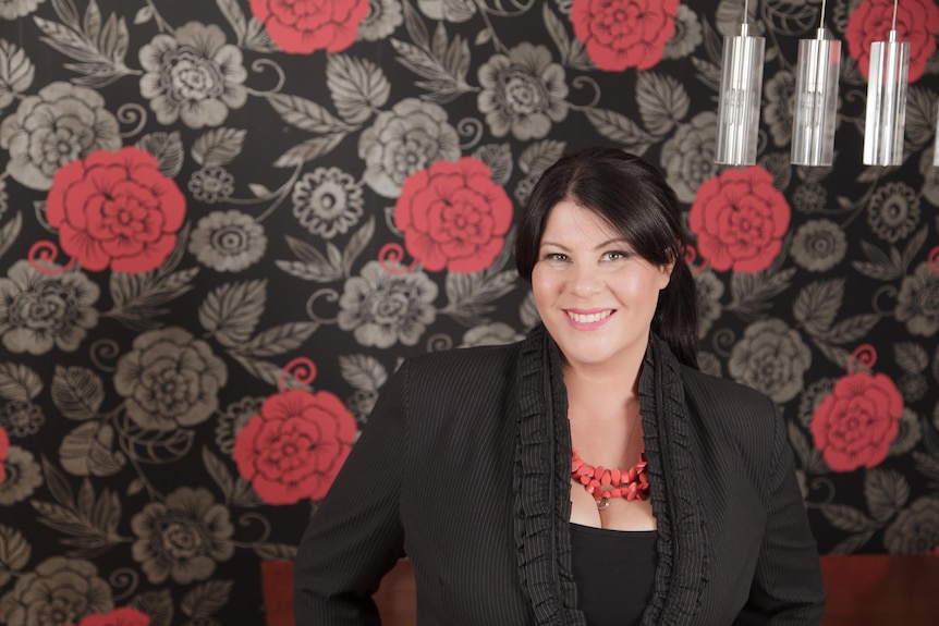 A woman in a black top in front of a wall with a floral wallpaper