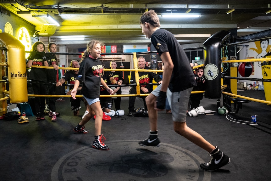 A young girl and a teenage boy circle one another in a boxing ring.