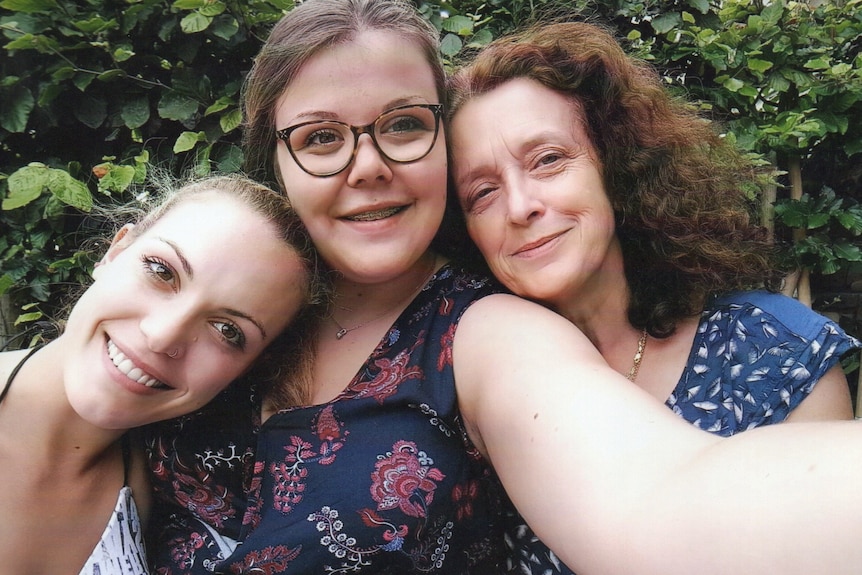 A young woman wearing thick-rimmed glasses holds arm out for 'selfie'. Her sister is on left and mother on her right, smiling