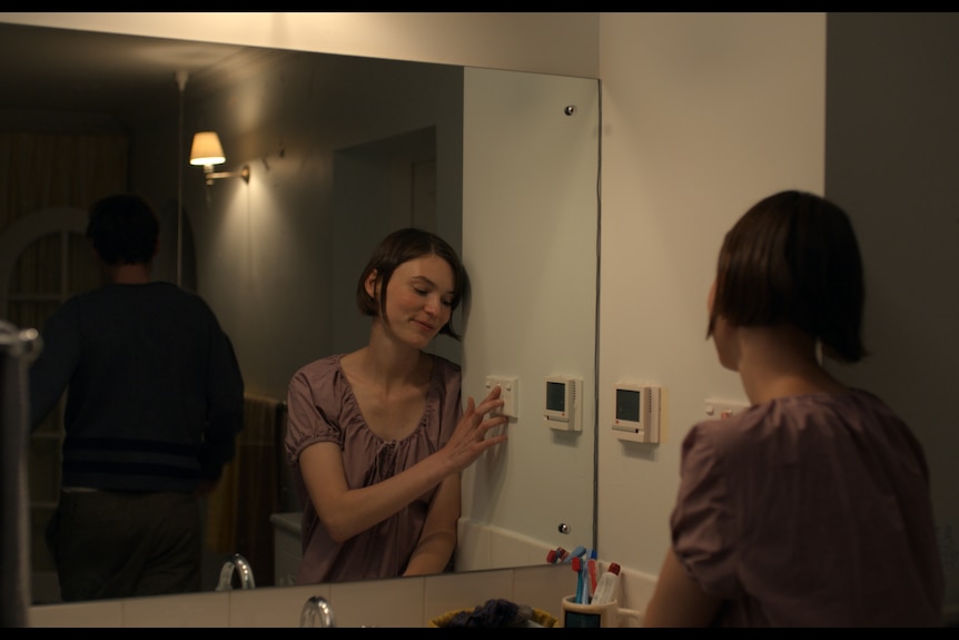 A woman stand in front of a mirror about to turn off the light while a man stands behind her