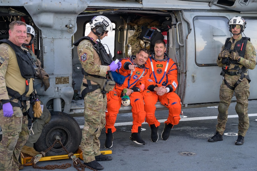 Two men in space suits smile as one of them points at the camera, they are sitting on a landed helicopter surrounded by soldiers