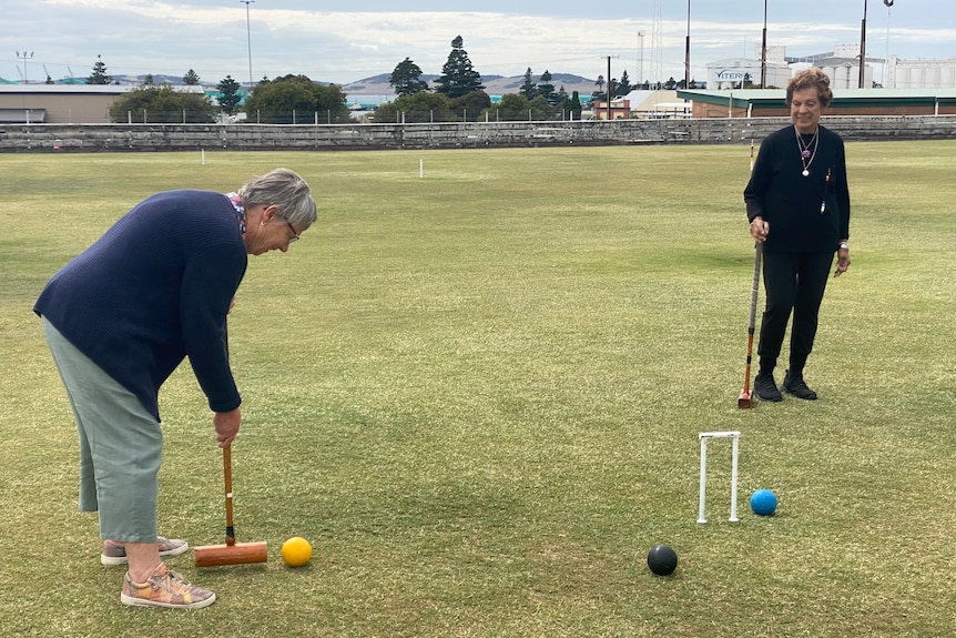 One woman having a croquet shot with a yellow ball, aiming for a hoop. Another woman watching on.