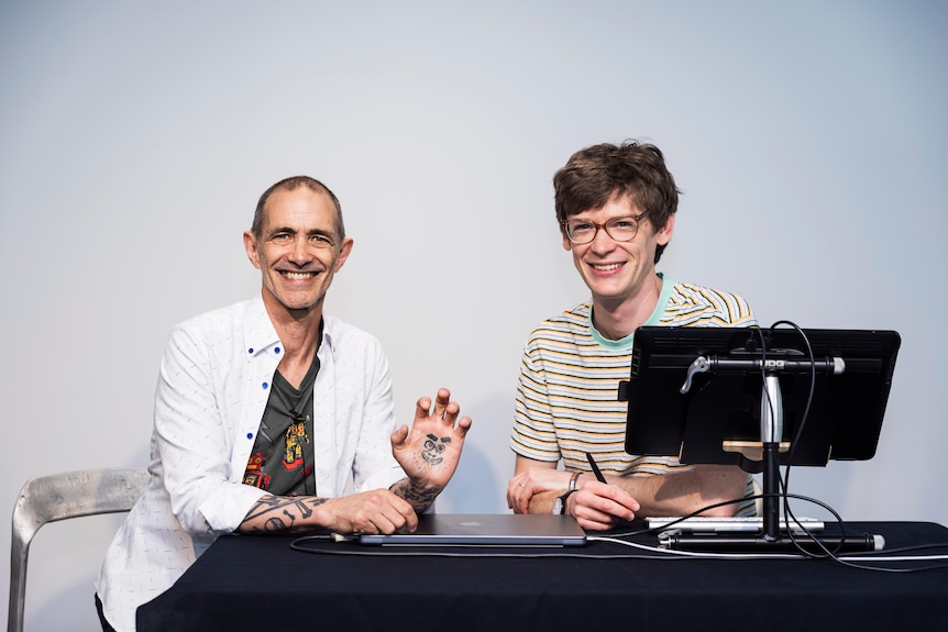 Andy Griffiths sits at a desk smiling, holding up a palm tattooed with a face, next to Bill Hope holding a pen 