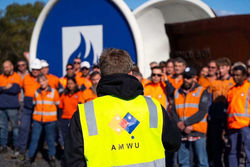 men and women in orange high vis outside a blue logo and a rusted sculpture