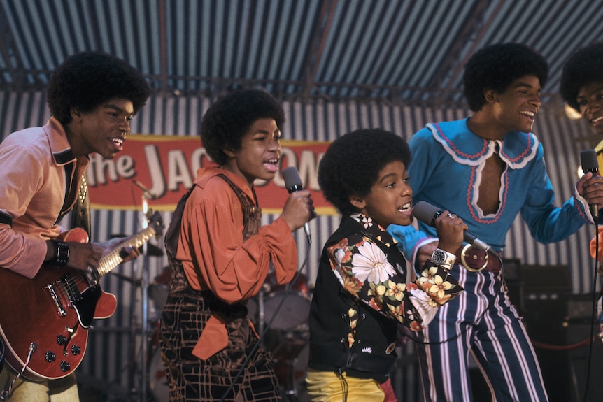 A group of five boys in 70s outfits sing on stage.