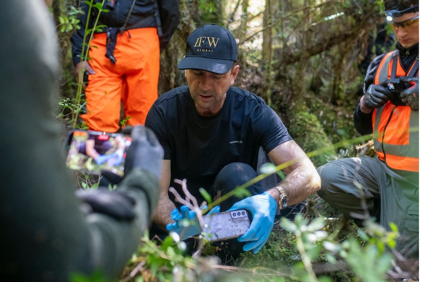 A man wearing black T-shirt, cap and blue gloves holds up a mobile phone just off the ground in a mossy forest