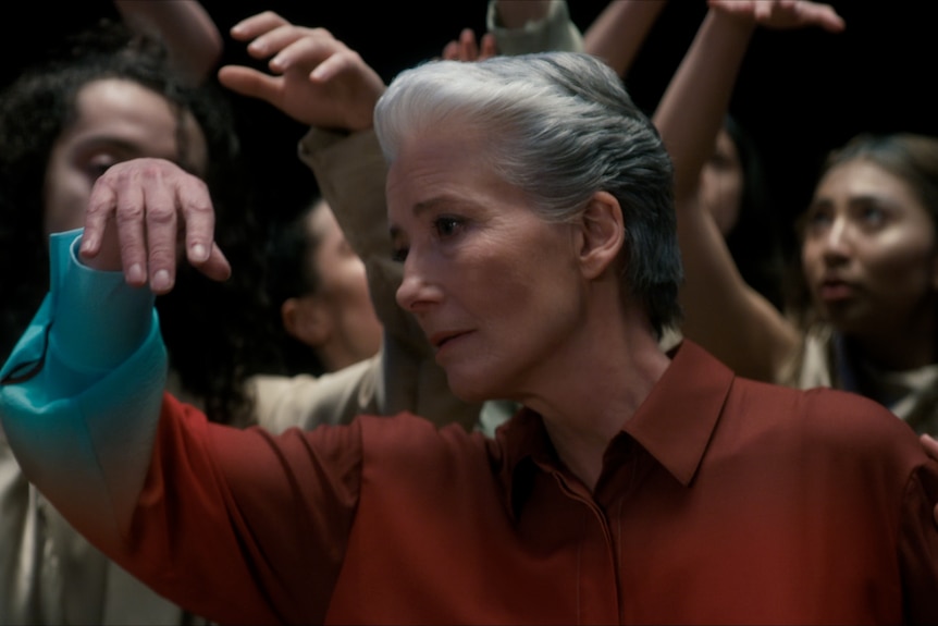 Emma Thompson dances in an orange shirt with one arm raised and a mournful expression. Dancers in beige surround her.