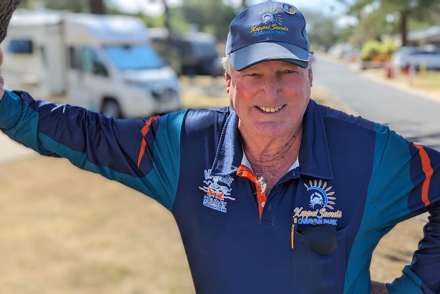 A man in a long sleeve polo leans against a tree while smiling in a caravan park.