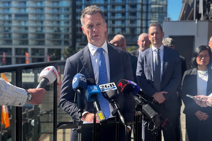 Mr Minns standing at a lecturn talking into a microphone with a big glass apartment building behind him.