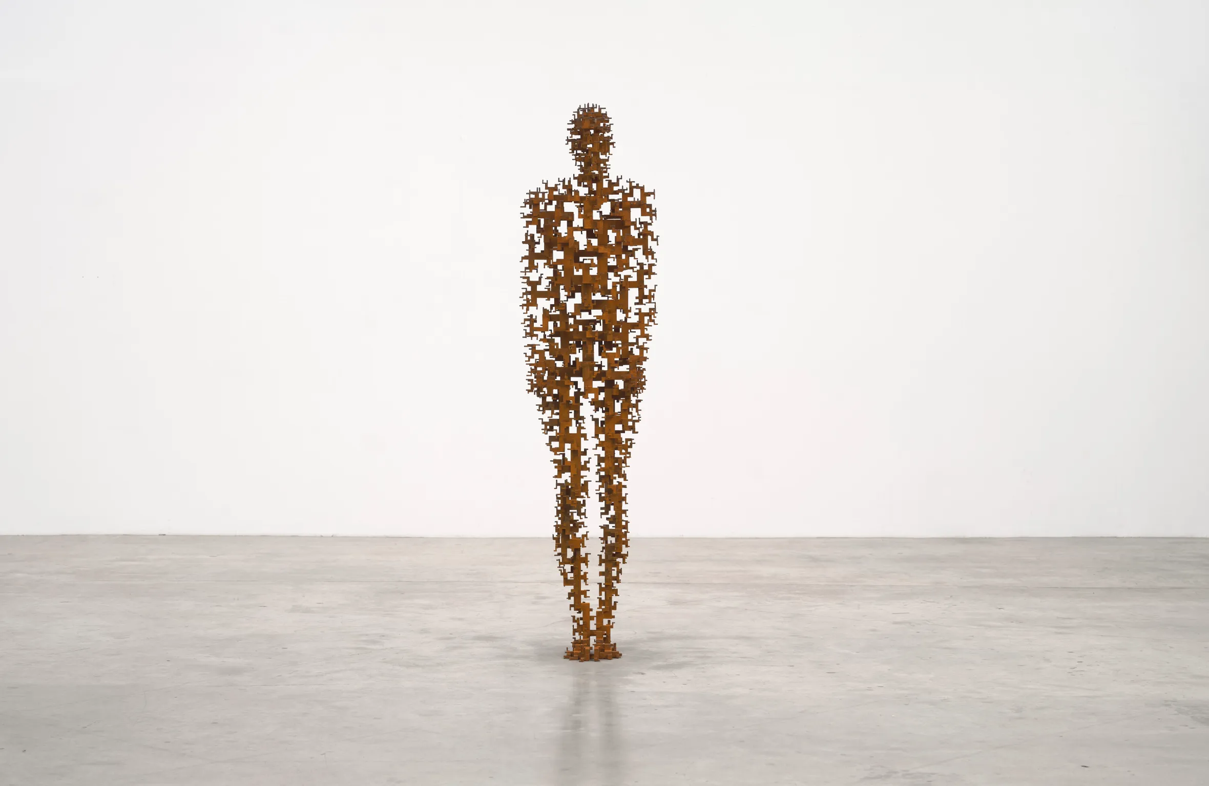 A life-sized, human-shaped sculpture made of interconnected, rusted iron forms, standing on a grey floor against a white wall.