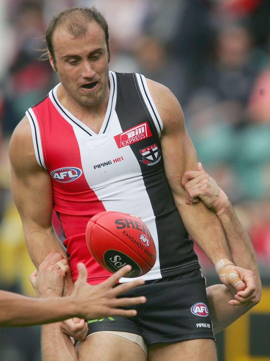 A St Kilda AFL player grimaces as his arms are held as he tries to mark the ball.