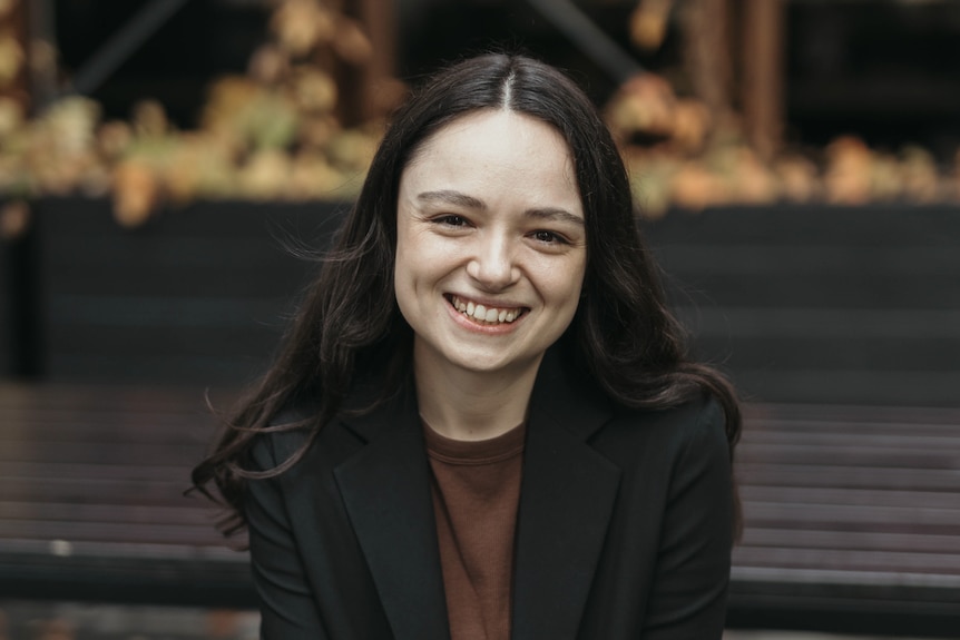 A young white woman with long dark hair sitting on a park bench and smiling