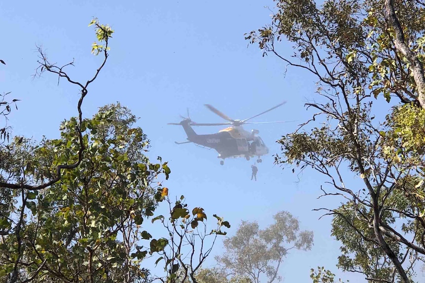 A blue and yellow helicopter flying with a stretcher being winched up beneath it framed by trees