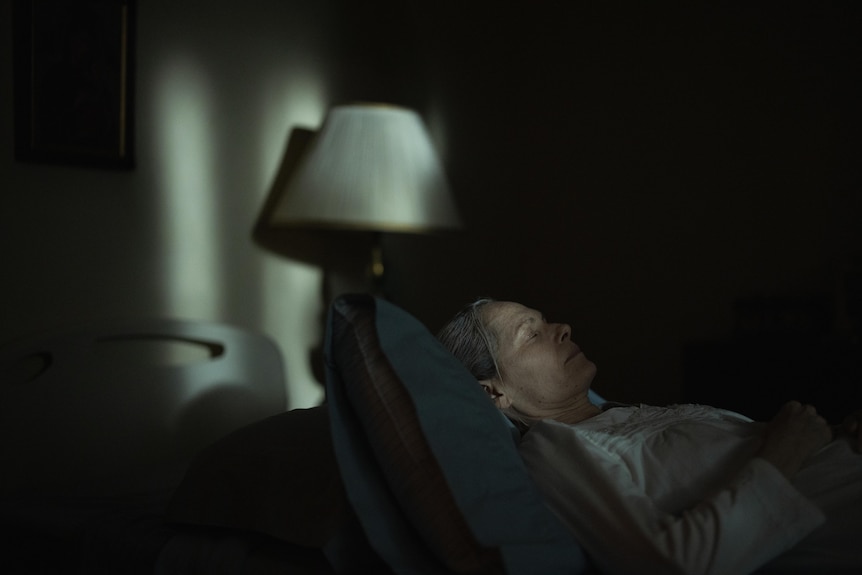 An elderly woman lies in a bed in a dark room