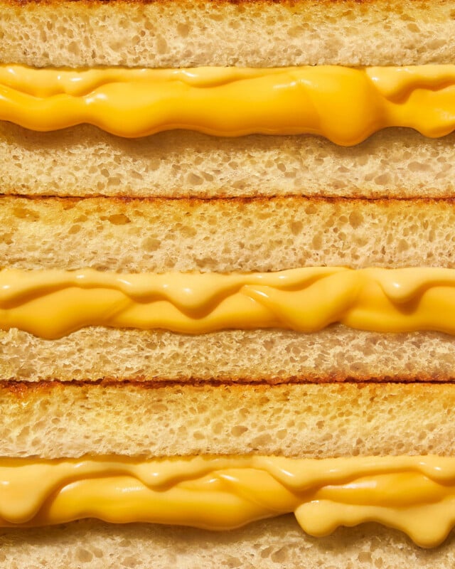 Close-up of three stacked grilled cheese sandwiches, showing golden brown toasted bread and melted yellow cheese oozing between the layers.