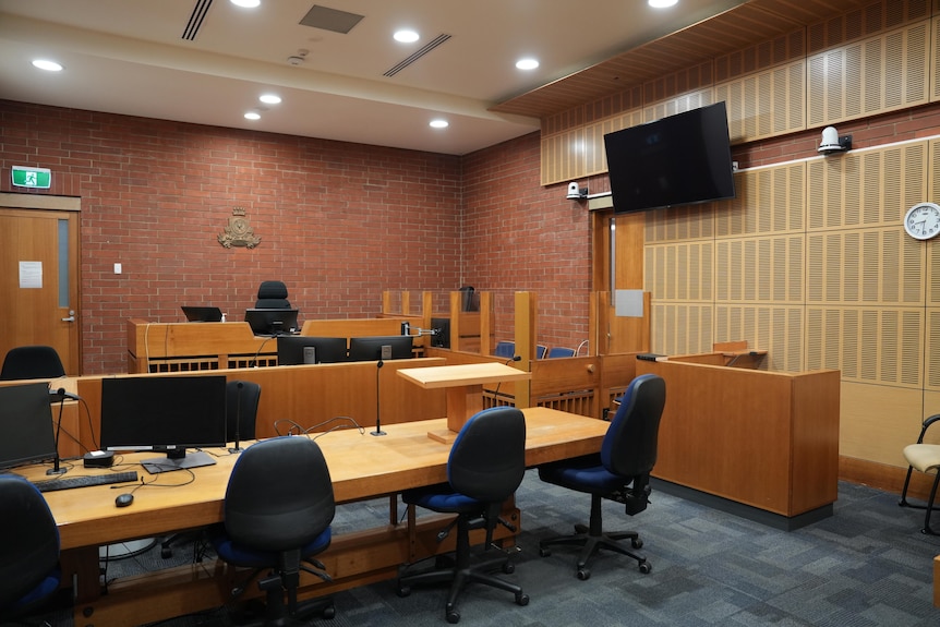 Empty desks and chairs facing the judge. A TV screens hangs on the wall above the defendent area fenced in by a half glass wall
