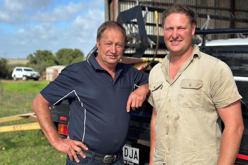Two men stand in front of a white uet with a green paddock behind them.