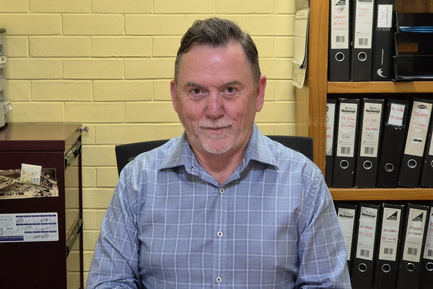 A man in a blue shirt seated at a desk with a yellow wall office behind him and a shelf full of folders.