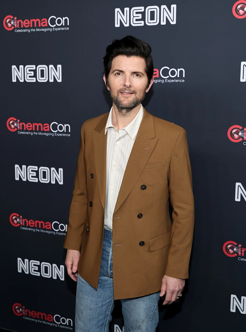 Adam Scott on a red carpet at CinemaCon wears a blazer over a shirt and jeans, standing in front of a step-and-repeat backdrop
