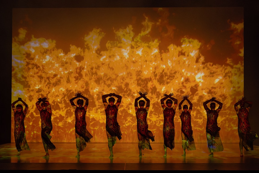 Nine dancers on stage with their arms above their heads, giant orange flames on the backdrop behind them
