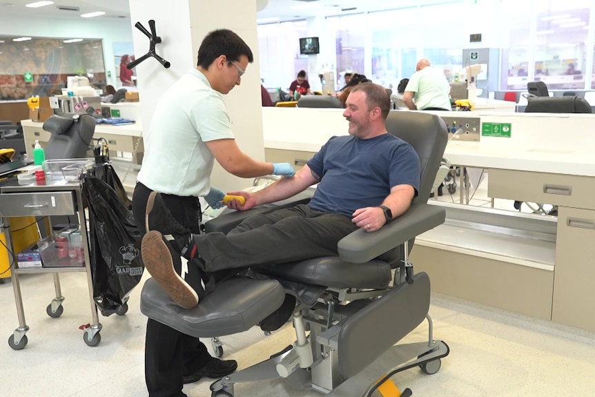 A blood donor centre staff swabs the inner elbow of a man sitting in a reclined chair 