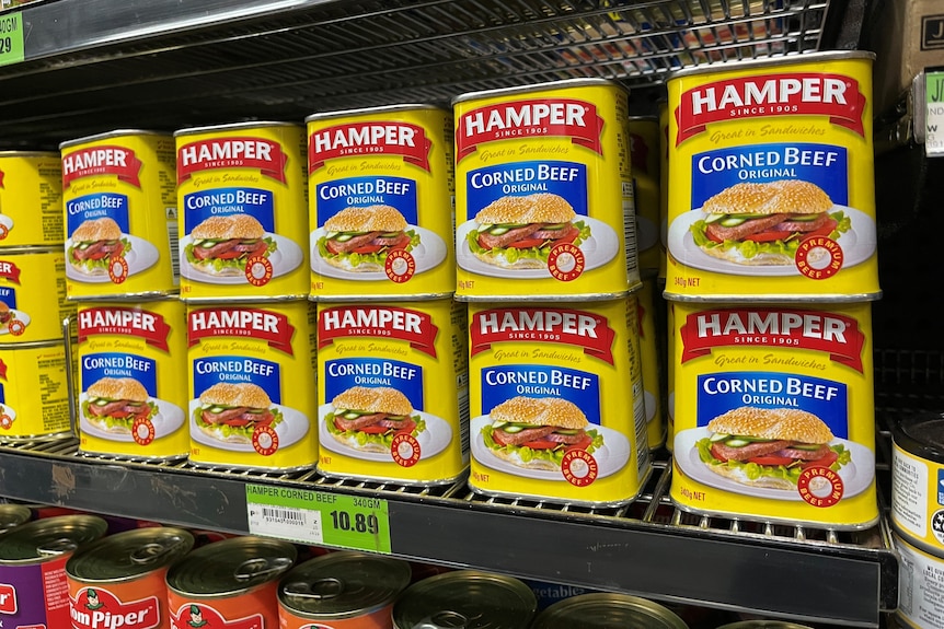 Small cans of corned beef are lined up on a supermarket shelf, the adjoining price tag reads: $10.89