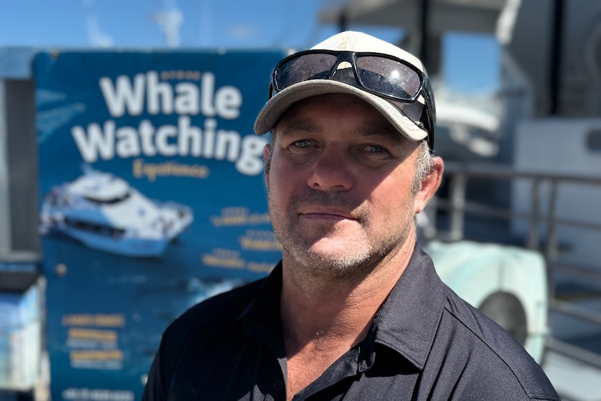 A man wearing a black polo shirt and cap standing in front of a whale watching sign