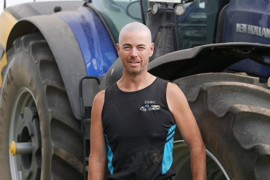 Cameron Dooner on a farm with his tractor behind him, smiling, has a shaved head, wears a sleevless singlet.