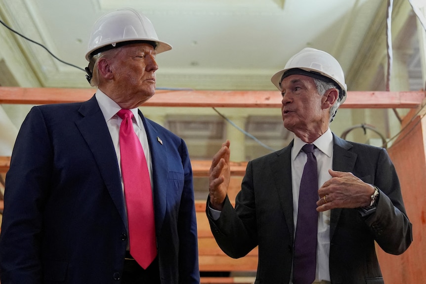 Donald Trump glares at a shorter man speaking to him while they both wear hard hats and suits on a construction site.