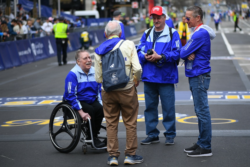 An older man in a wheelchair speaks with a group of marathon officials.