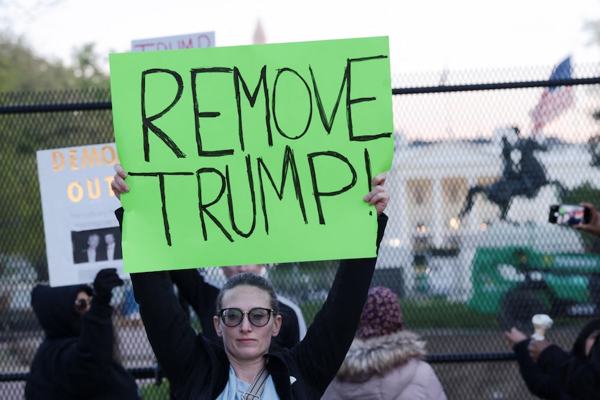 A woman holds up a sign that says "REMOVE TRUMP" in front of the White House.