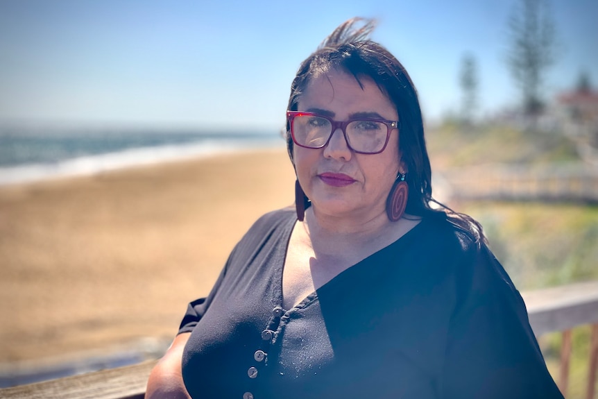 A woman with red glasses, orange and brown circular earrings wearing a black top leans her elbow on a wooden post at the beach.