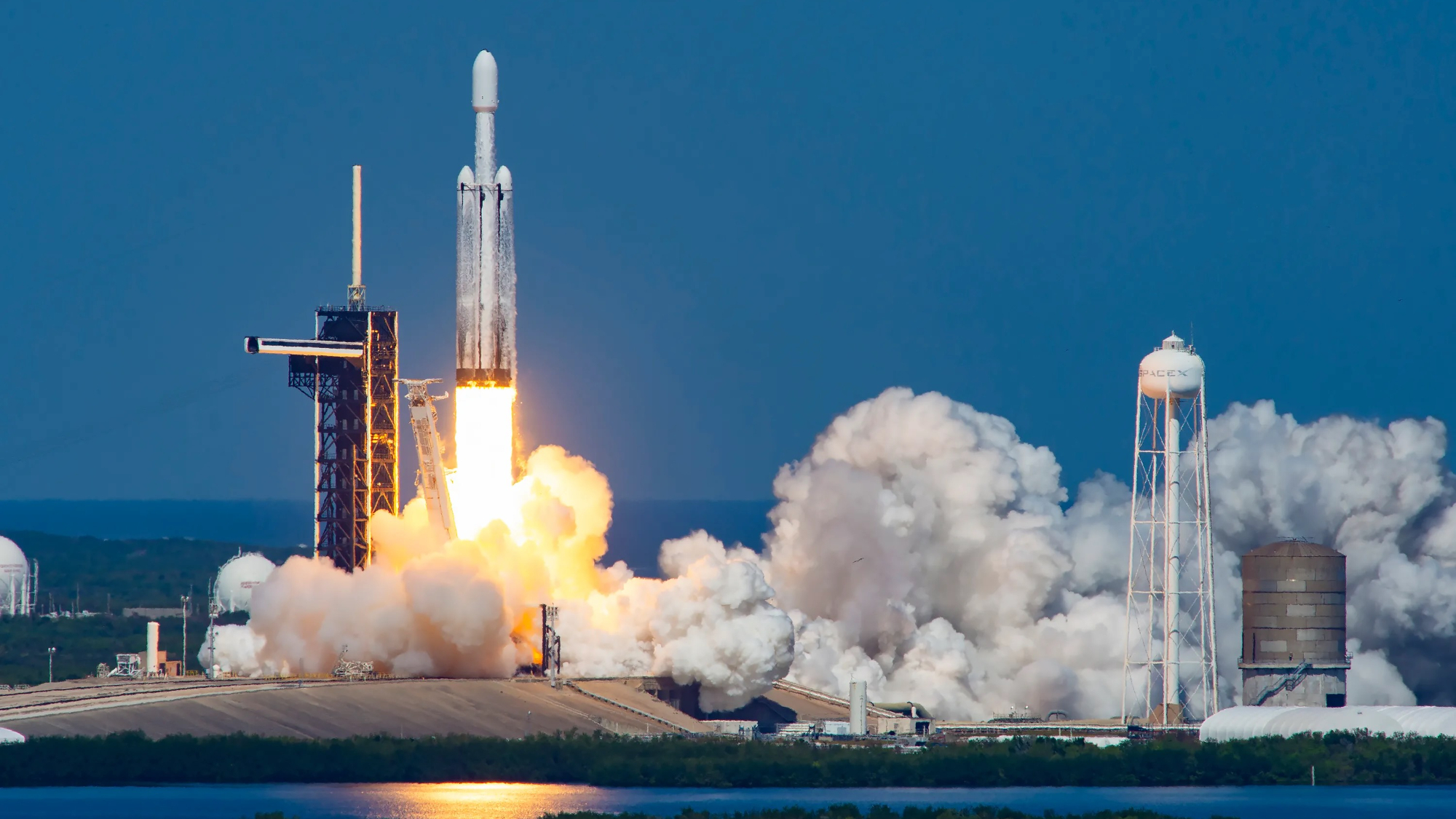 a big white rocket launches into a blue sky from a seaside pad