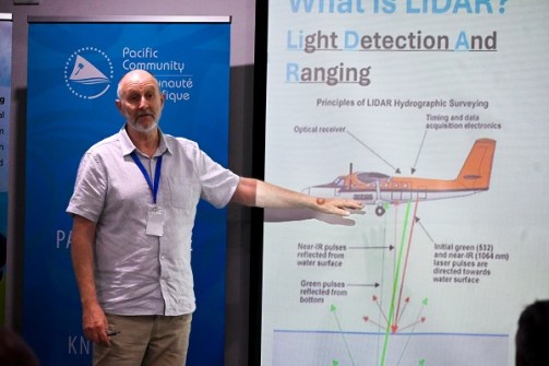 A man standing and pointing to an image of an aircraft display on a large screen, during a presentation on aerial LiDAR scanning