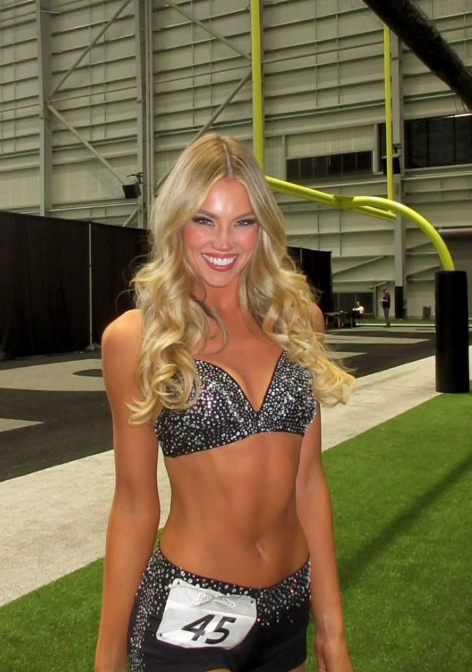 A female cheerleader with long blonde hair and wearing a sparkly black top and shorts with the number 45 smiles at the camera on a football field.