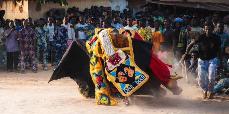A performer in a colorful, beaded masquerade costume dances energetically in the center of a dusty outdoor area, surrounded by a crowd of onlookers, many dressed in vibrant patterned clothing.