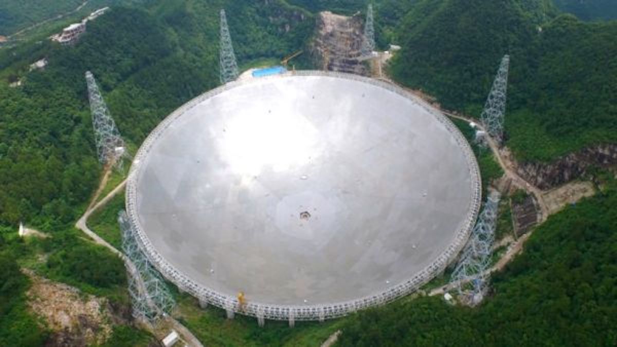aerial photo of a huge, silvery radio telescope dish surrounded by tree-covered mountains