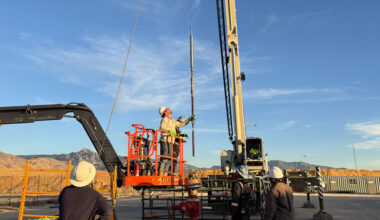 A worker on a scissor lift holds a long pole connected to a crane. Two workers are visible in the foreground.