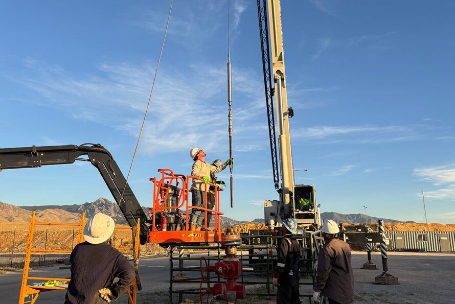 A worker on a scissor lift holds a long pole connected to a crane. Two workers are visible in the foreground.