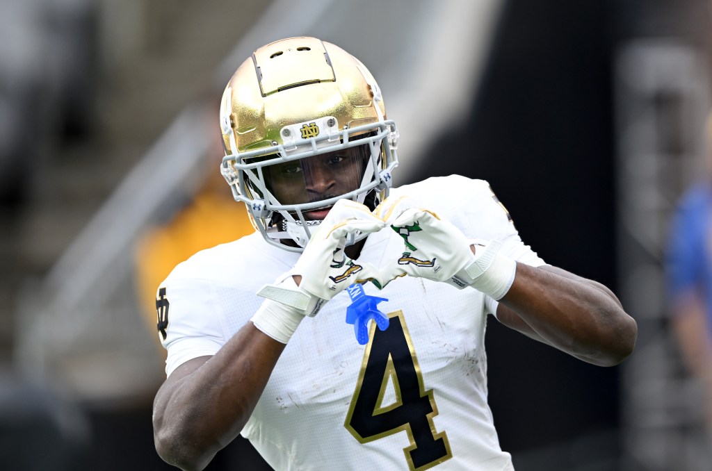 A Notre Dame football player in a white uniform and gold helmet makes a heart shape with his hands.