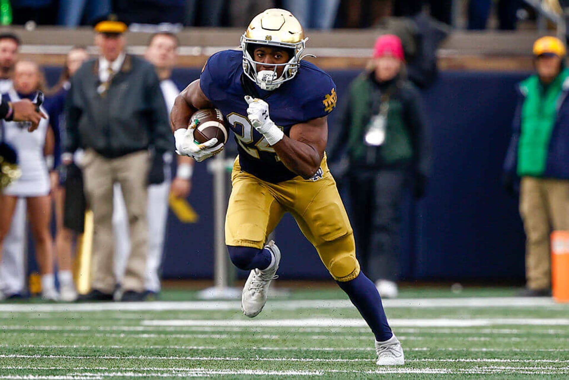 Former Notre Dame running back Jadarian Price, clad in a navy blue Fighting Irish jersey with gold pants and helmet, strides in the open field with the ball tucked into his right side.